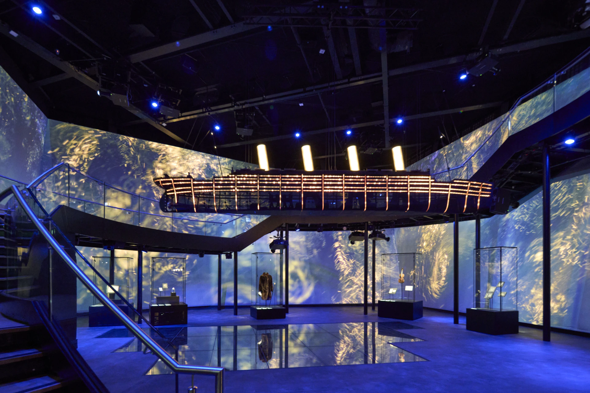 A museum exhibit featuring a suspended wooden boat under dramatic lighting, with glass displays and digital projections around.