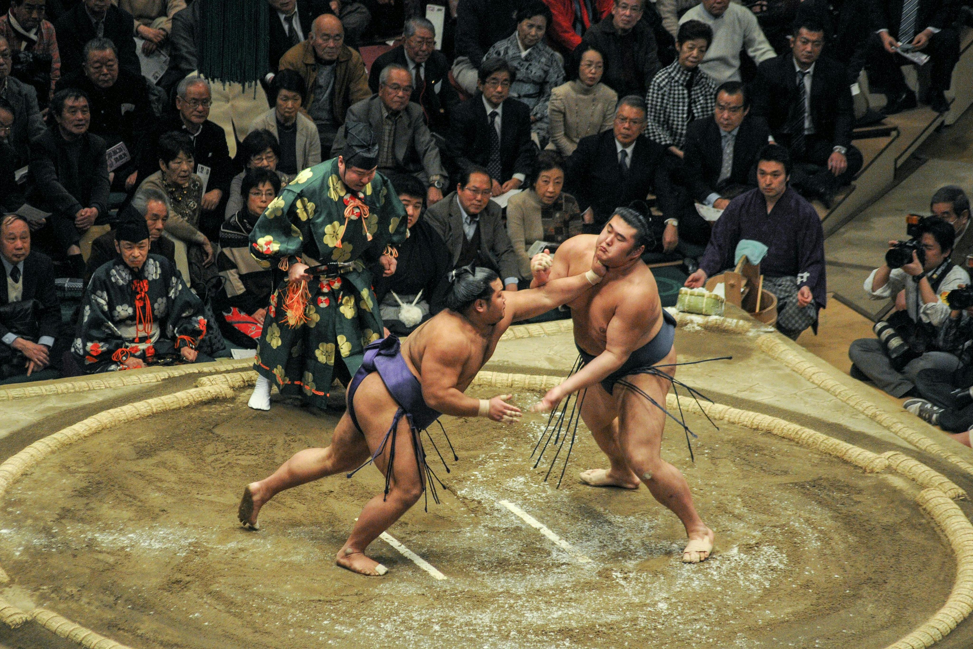 Two sumo wrestlers competing in a ring surrounded by spectators, with a referee standing nearby.