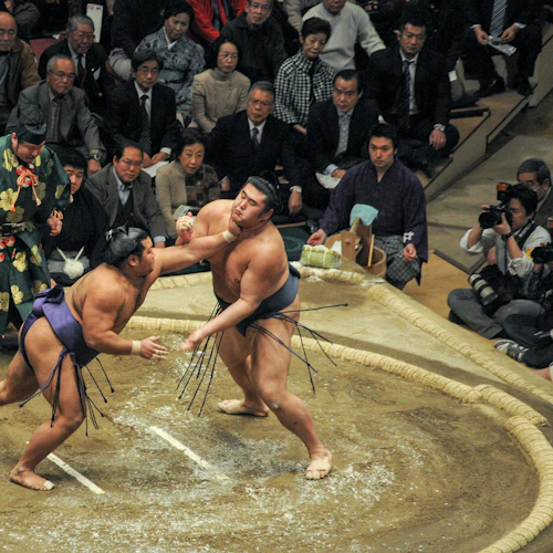 Two sumo wrestlers competing in a ring surrounded by spectators, with a referee standing nearby.