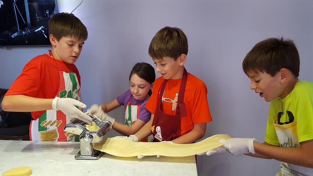 Τρία παιδιά φτιάχνουν ζυμαρικά μαζί, χρησιμοποιώντας μια μηχανή ζυμαρικών σε έναν πάγκο. Δύο αγόρια και ένα κορίτσι φορούν ποδιές και γάντια.