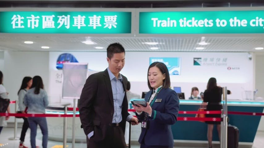 スーツ姿の男性と制服姿のスタッフが、列車の切符売り場の前で、英語と中国語の看板でやり取りしている。