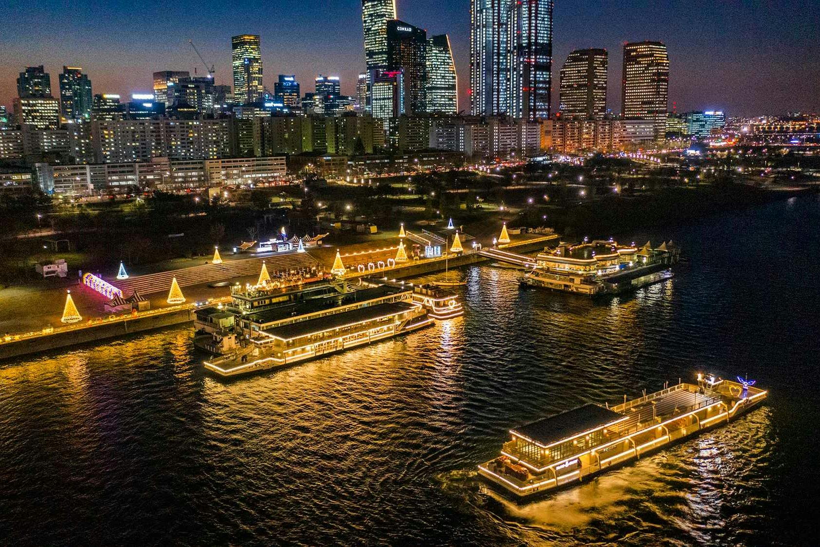 Un paisaje urbano nocturno con transbordadores iluminados en un río, rodeados de edificios altos y muelles iluminados.