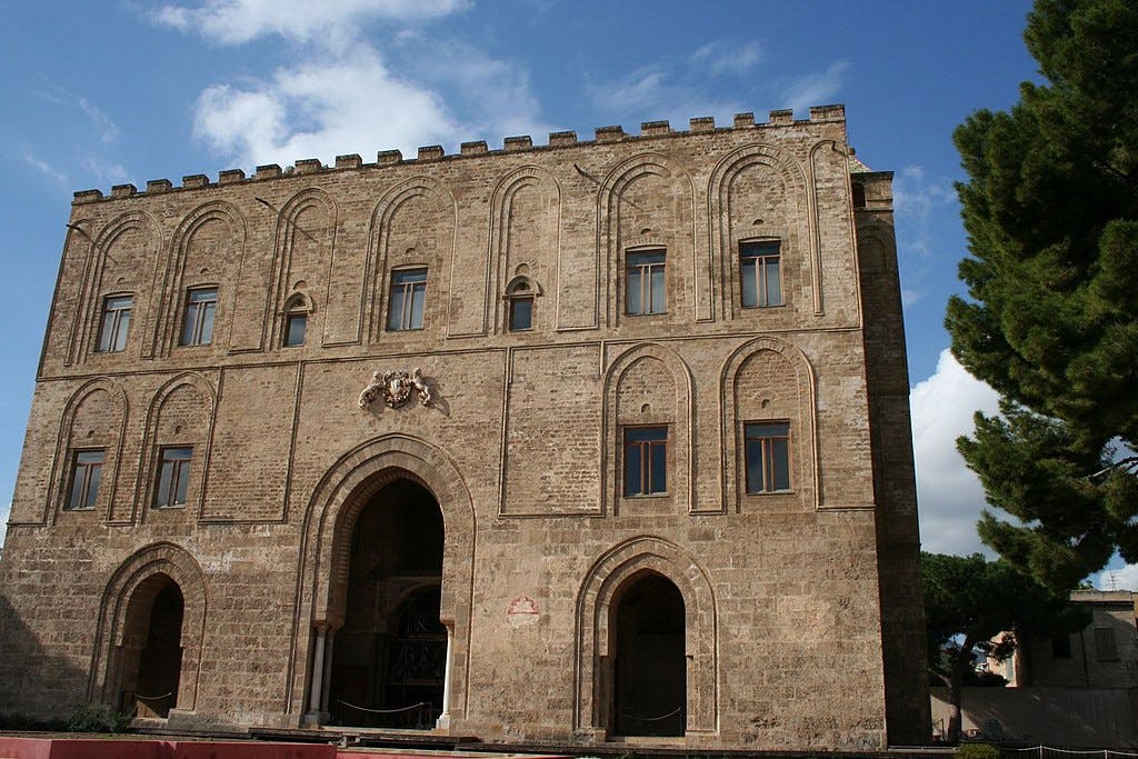 Bâtiment historique en pierre avec des fenêtres et des portes cintrées, présentant des sculptures complexes et un style architectural médiéval. Arbres sur la droite.