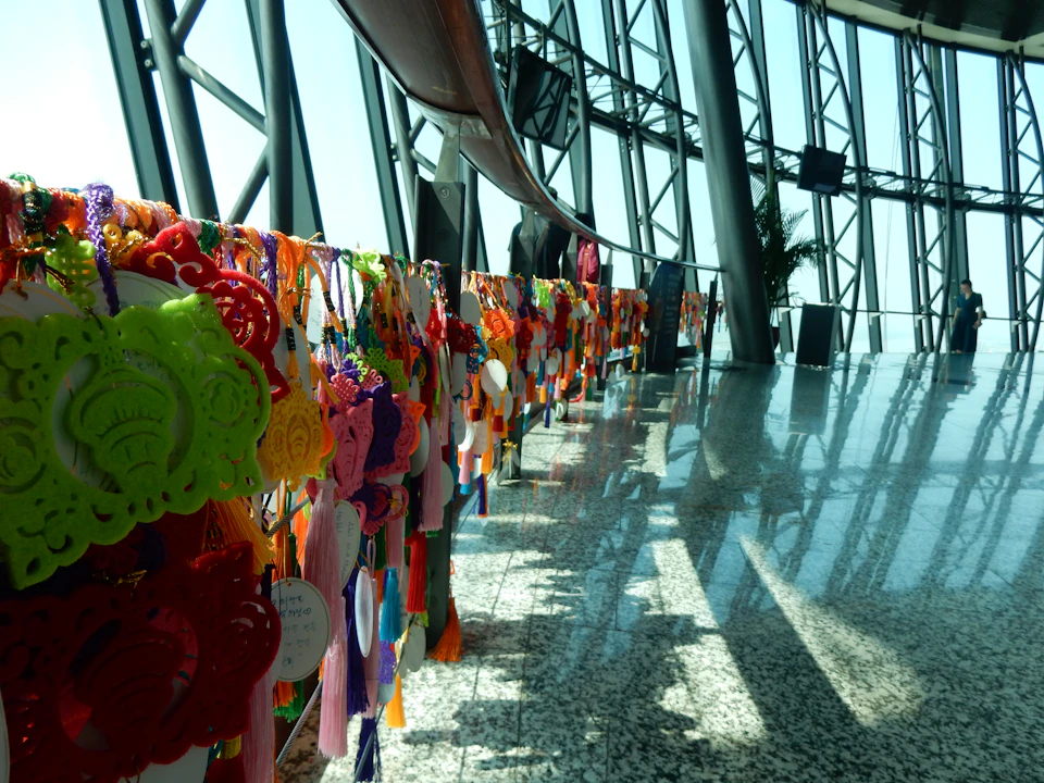 Colorful hanging decorations line a glass-walled corridor with sunlight casting shadows on the granite floor.