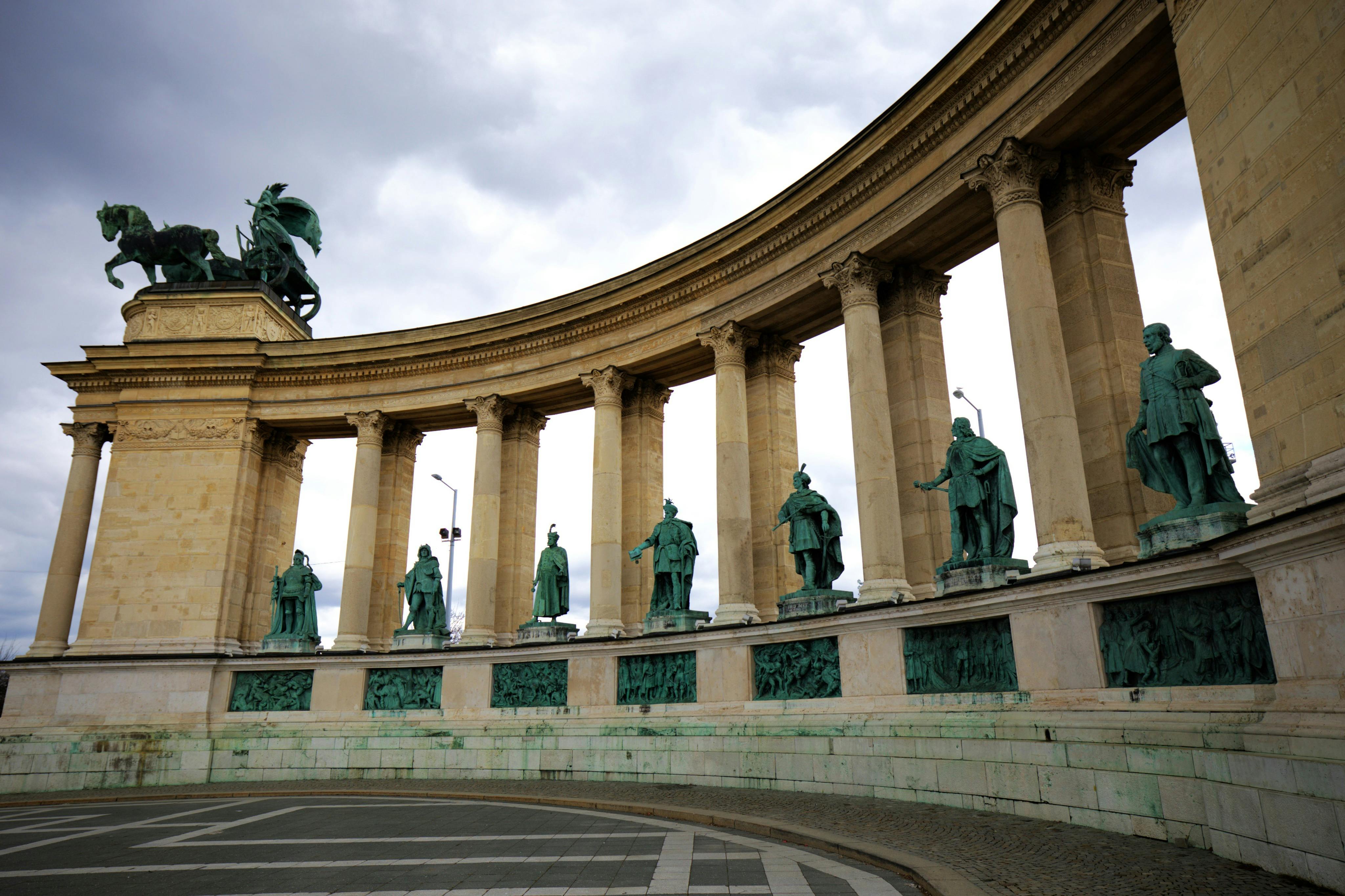 A semi-circular colonnade with statues of historical figures under a cloudy sky, part of a monumental architectural structure.