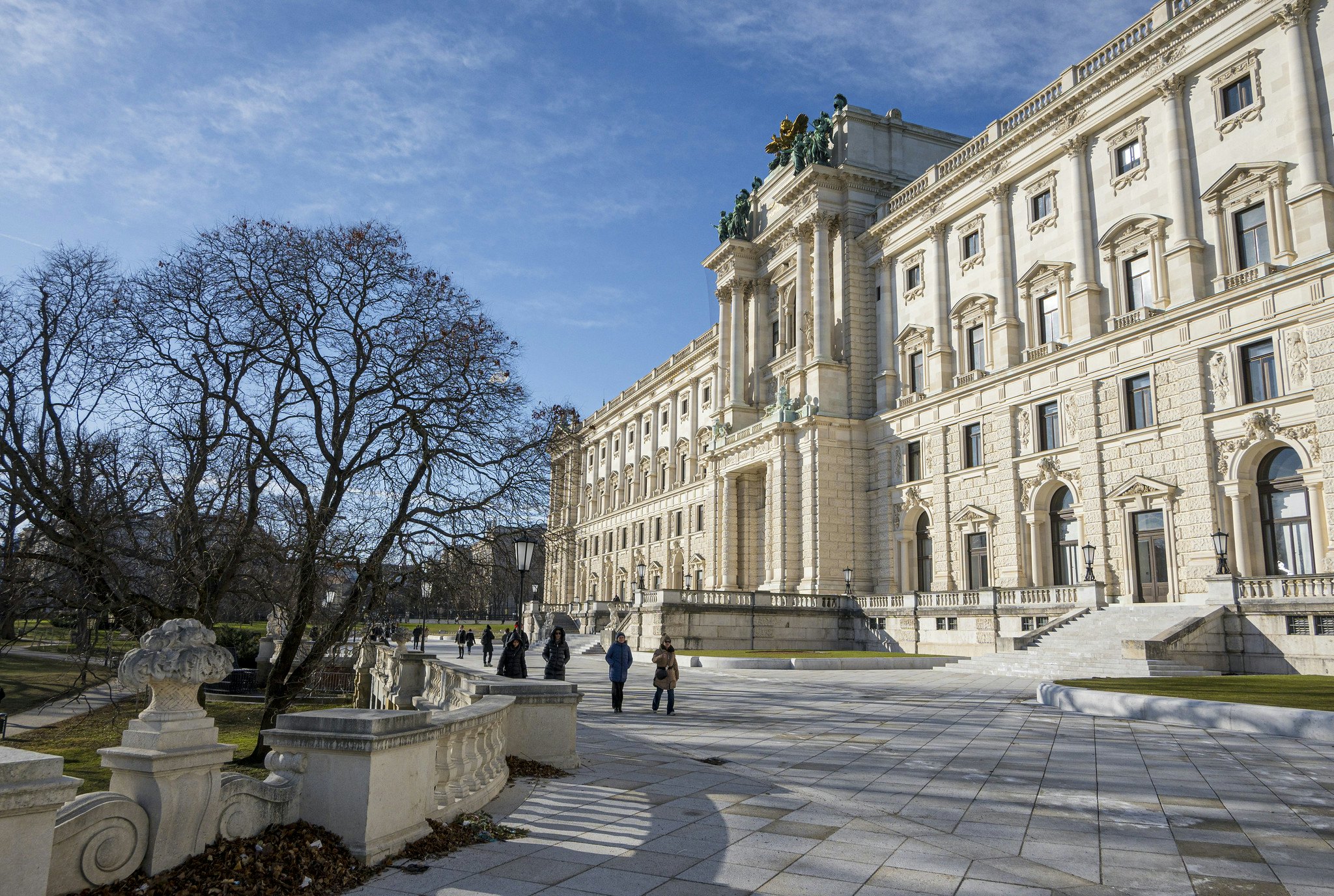 Lidé kráčející kolem velké historické budovy s ozdobnou architekturou, obklopeni nádvořím, stromy a jasně modrou oblohou.