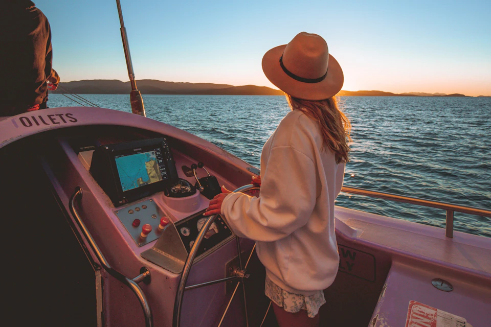 Person in a hat steering a boat at sunset, with a view of water and distant mountains.