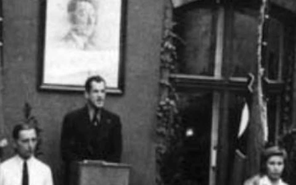 A group of people stands in front of a building with a man at a podium, a Nazi flag, and a banner with German text above.