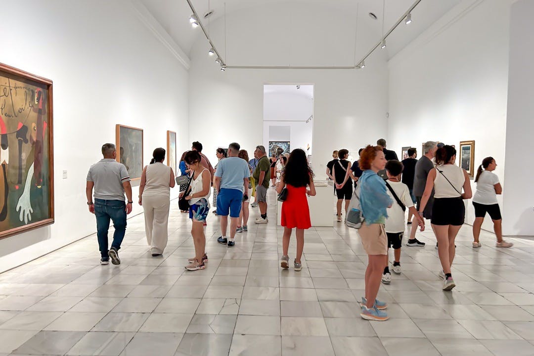 Folk, der går og observerer kunstværker i et lyst, hvidvægget galleri med malerier hængt på væggene.