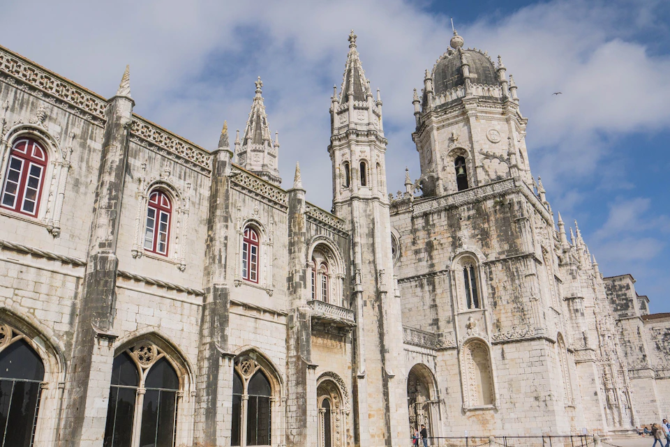 A historic stone building with ornate Gothic architecture, featuring tall spires and arched windows under a partly cloudy sky.