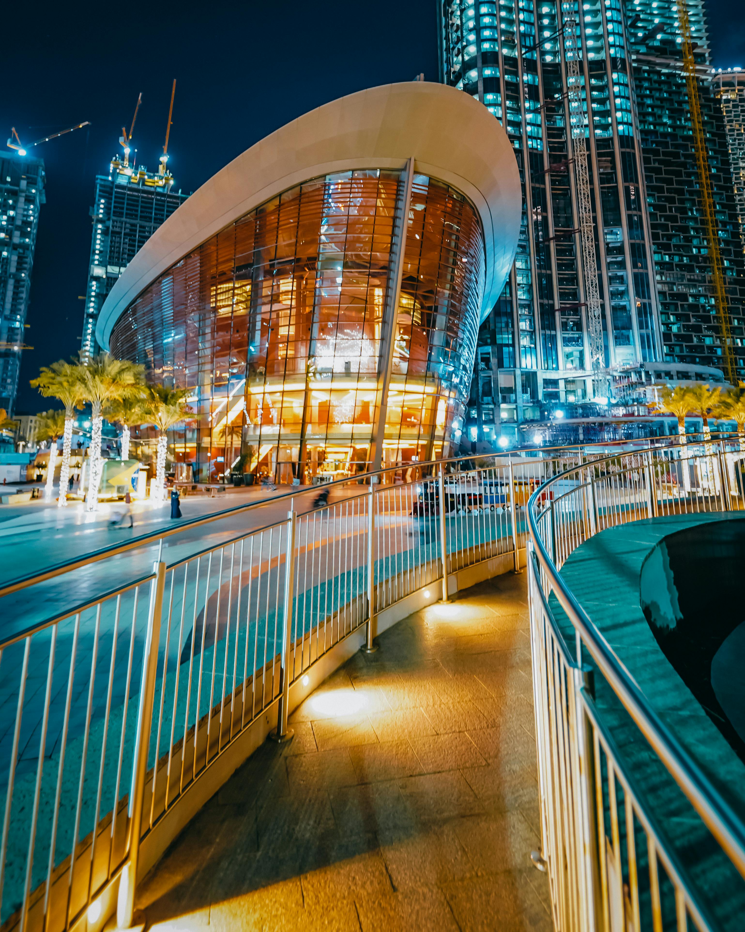 Curved pathway with rails leading to a modern, glass-walled building illuminated at night, surrounded by other tall structures.