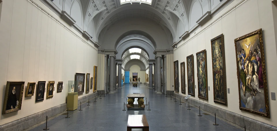 An art gallery hall with high arched ceilings, framed paintings on both walls, and sculptures displayed on center tables.