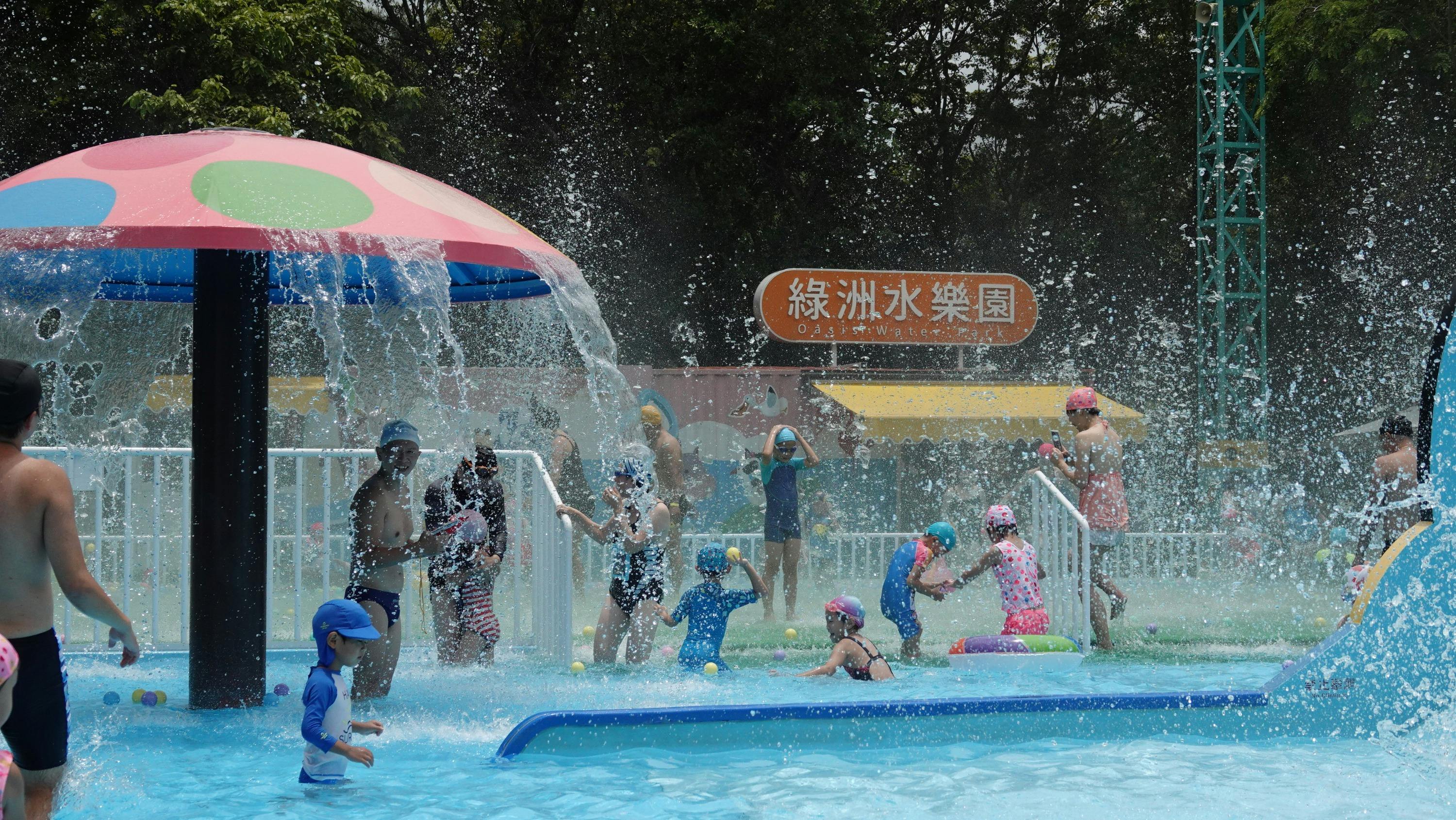 Children and adults playing under water sprays in a colorful outdoor water park with a sign in the background.