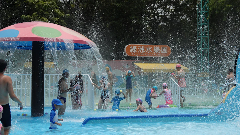 Children and adults enjoying a water play area with fountains and a colorful canopy, surrounded by trees and a park sign.