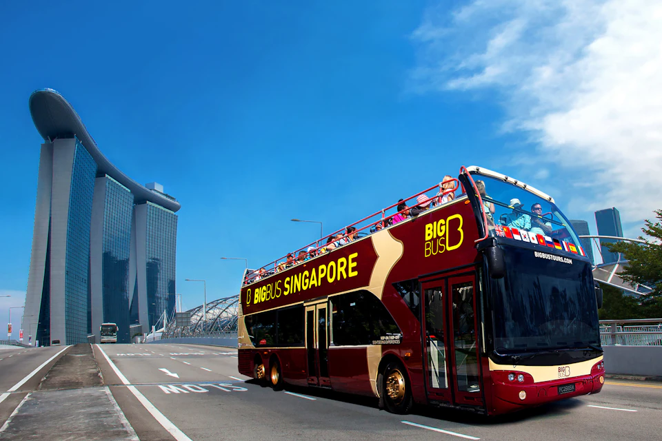 People on an open-top bus tour in a city with tall modern buildings, lush greenery, and several cars on the street.