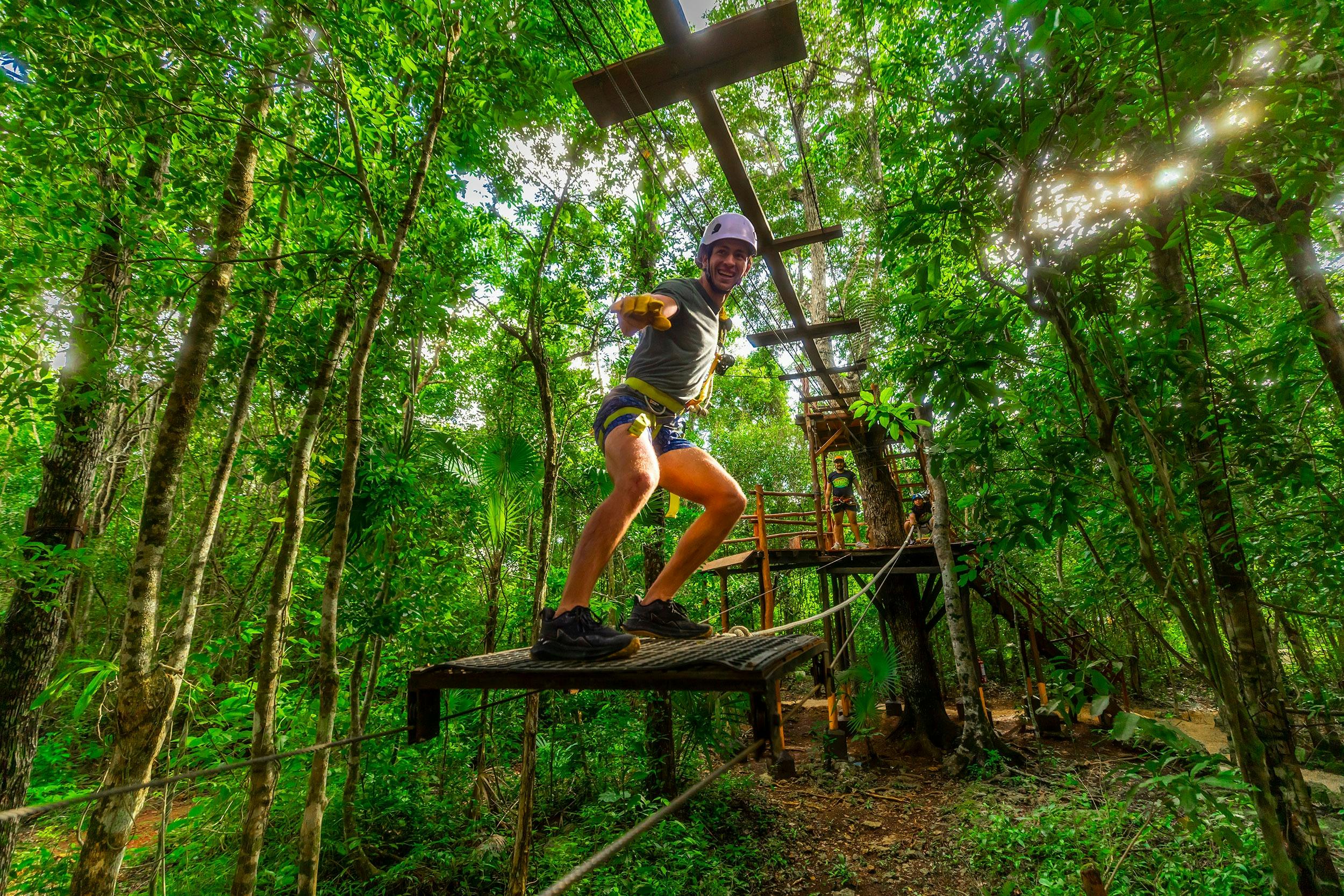 A person balancing on a rope bridge in a forest adventure park, wearing a helmet and harness, surrounded by lush greenery.