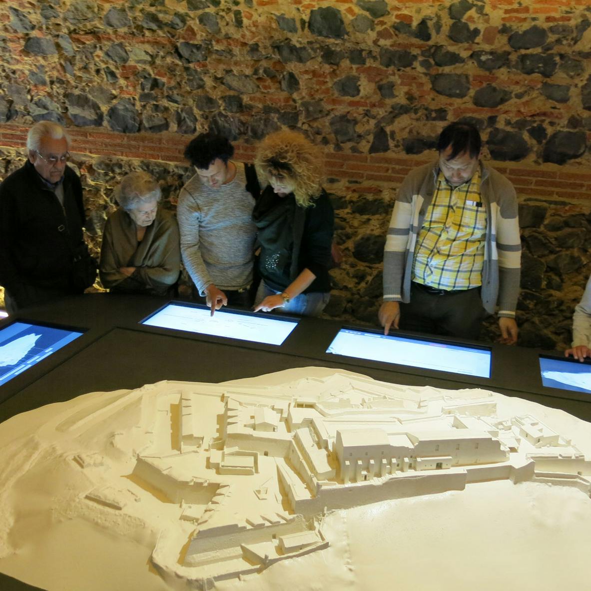 Five people interact with touchscreen displays around a detailed architectural model, with a stone wall in the background.