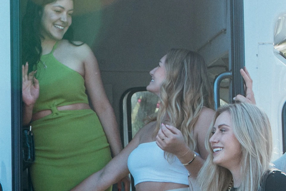 Five women stand and smile near the open door of a white tour bus with "Pam Tours" and a phone number displayed.