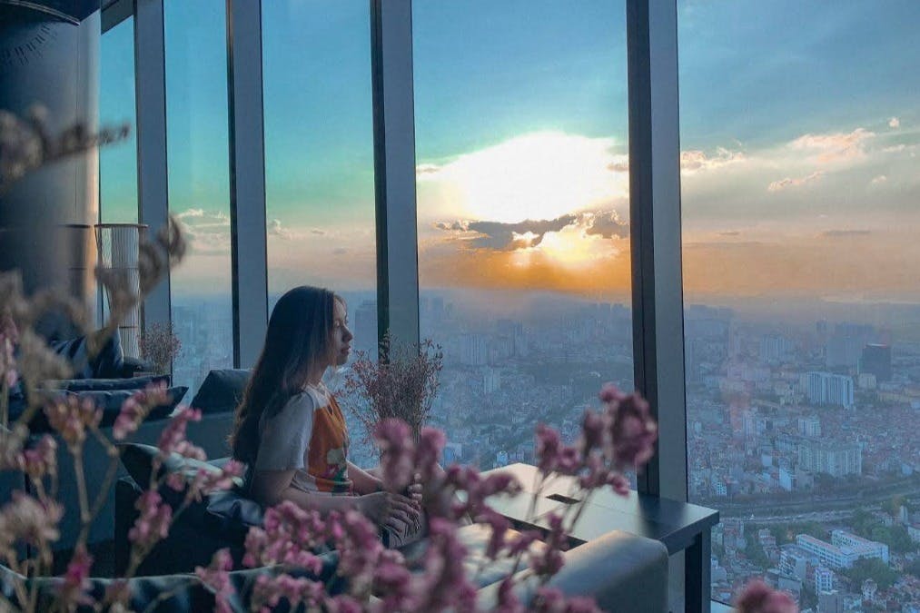Person sitting by large windows, overlooking a cityscape at sunset. Pink flowers are in the foreground.