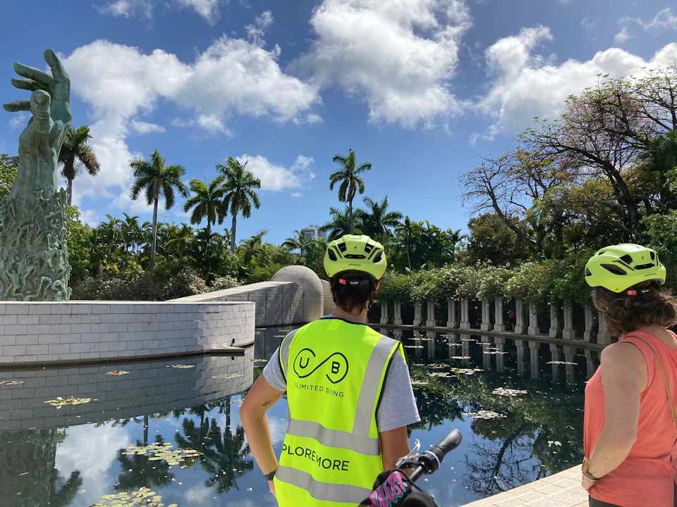 Person wearing helmet and "Unlimited Biking" vest stands by a serene pond with lily pads, surrounding palm trees, and white columns.