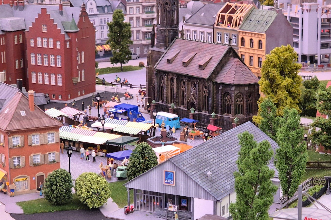 A miniature model scene depicting a marketplace with stalls, people shopping, a historic church, and an Aldi store in the foreground.