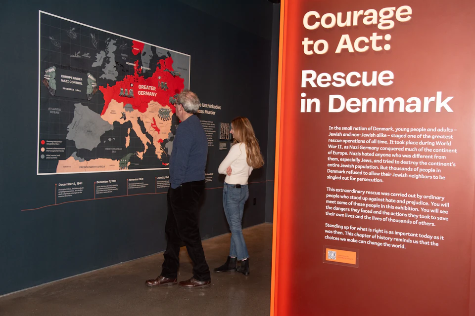 Two people view an exhibit featuring a map and text about Denmark's rescue efforts during WWII, with a large red panel displaying the title "Courage to Act."