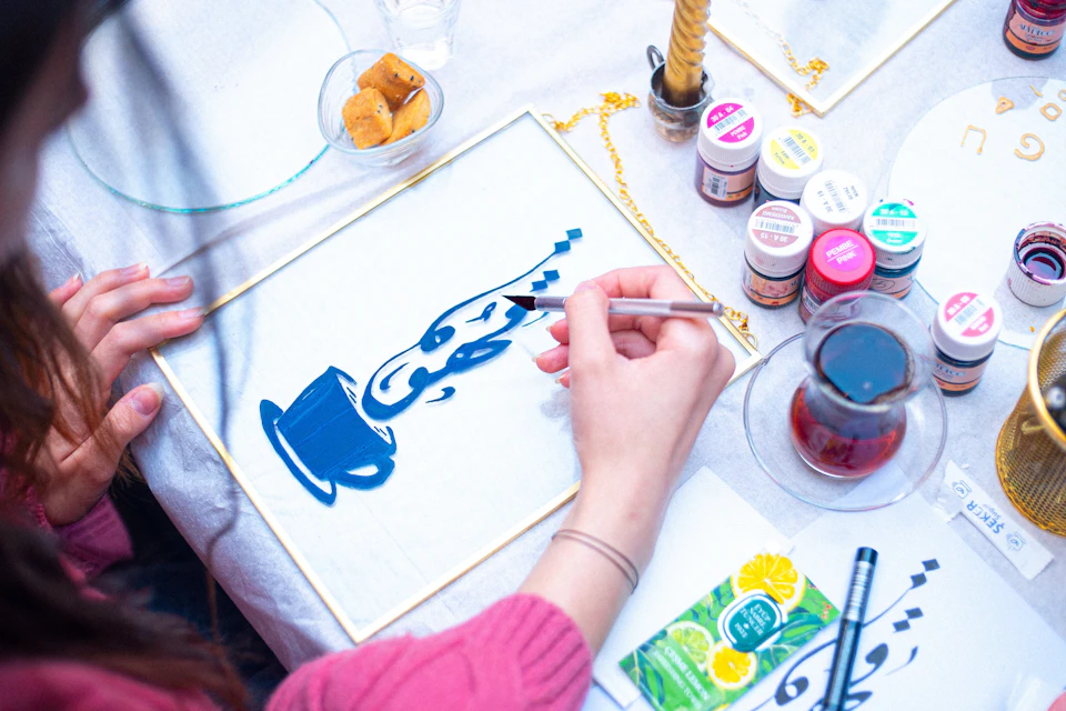 A person painting a blue cup and steam design on a canvas, with art supplies and a glass of tea on the table.