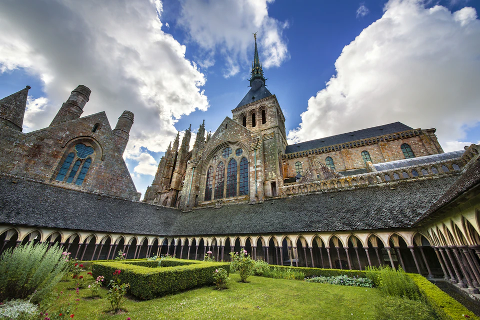 Stone building with a spire and arched windows, surrounded by a courtyard garden with trimmed hedges and blooming flowers under a partly cloudy sky.