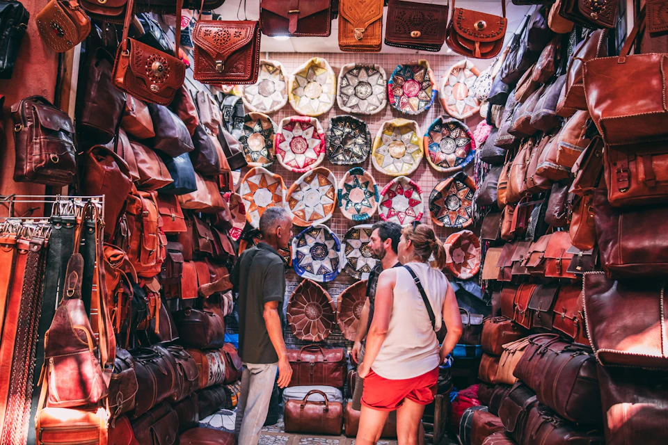 Three people browse a market stall filled with leather bags and colorful woven products displayed on the walls.