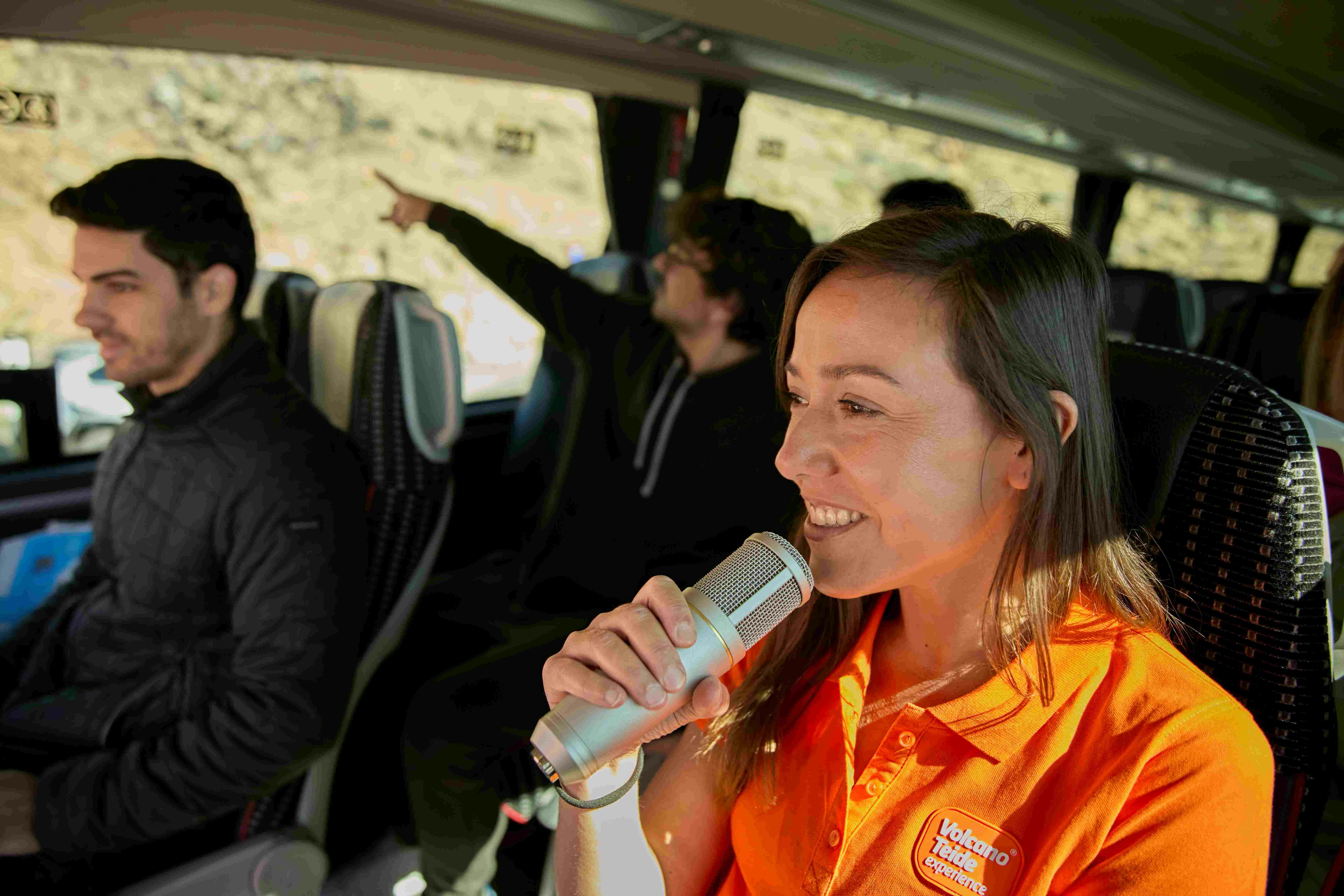 A woman in an orange shirt speaks into a microphone on a bus, with two seated passengers, one of whom is pointing.