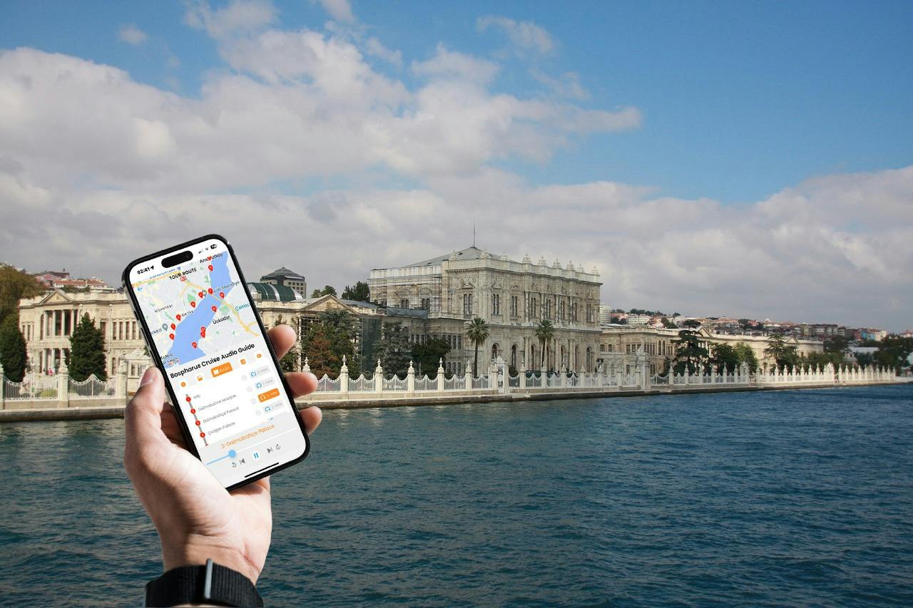 A hand holding a smartphone displaying a map app, with a historic waterfront building and blue sky in the background.
