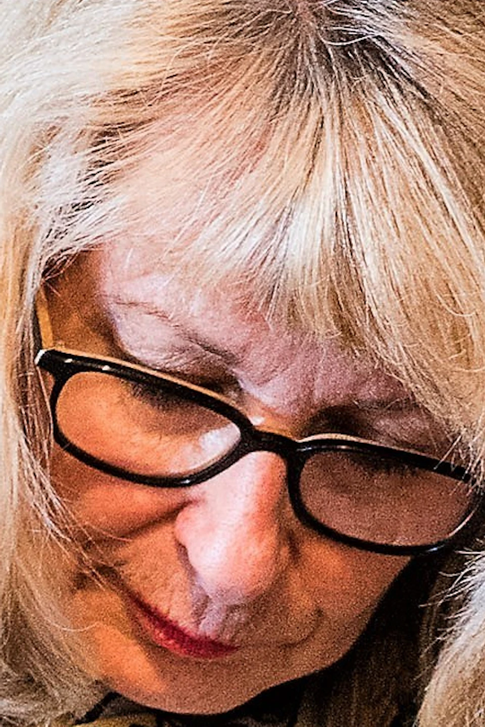A woman with blonde hair and glasses works with clay in an art studio, surrounded by artwork and a window in the background.