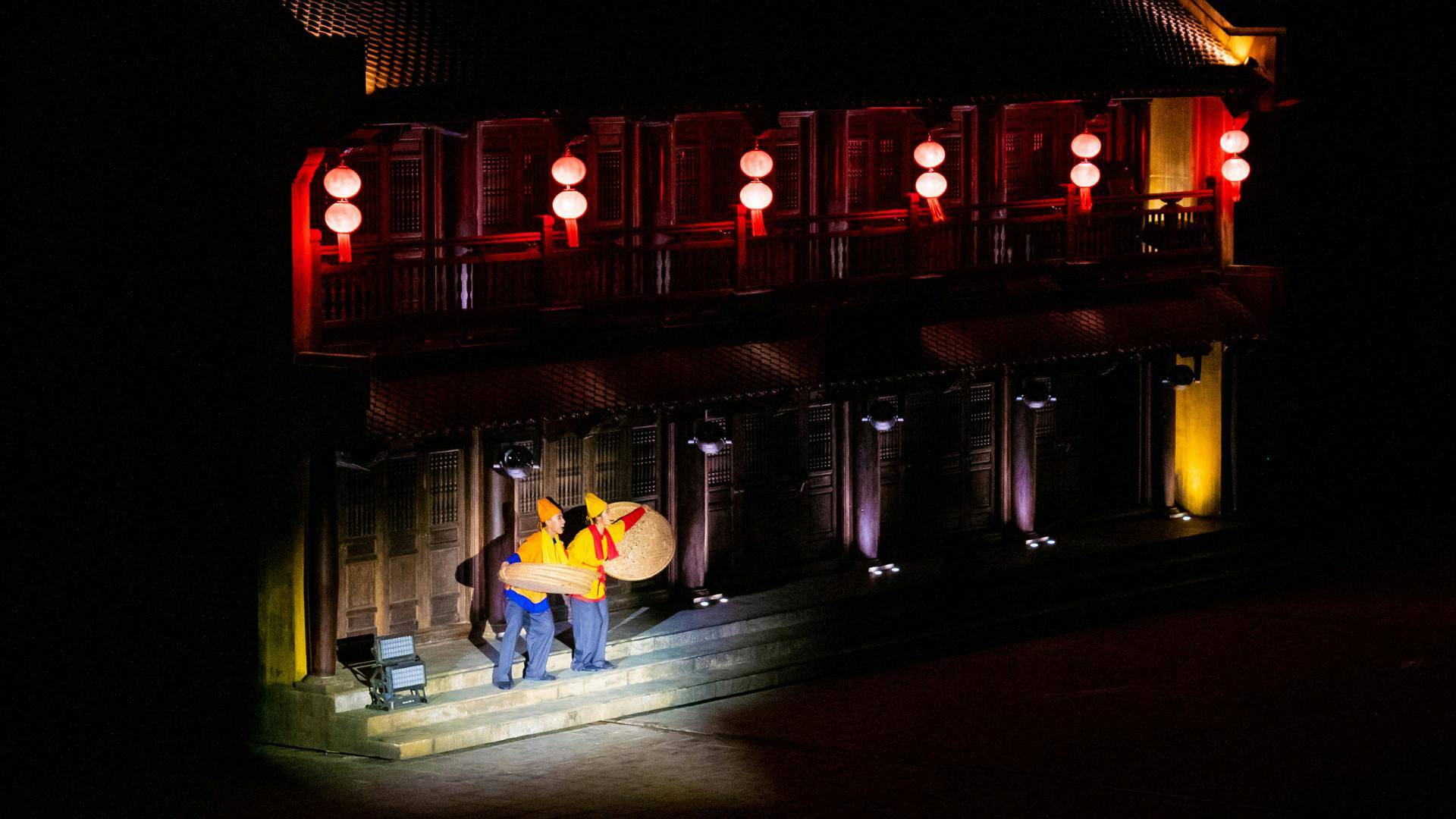 Two performers in colorful costumes hold a large prop on a stage with red-lit traditional architecture in the background.