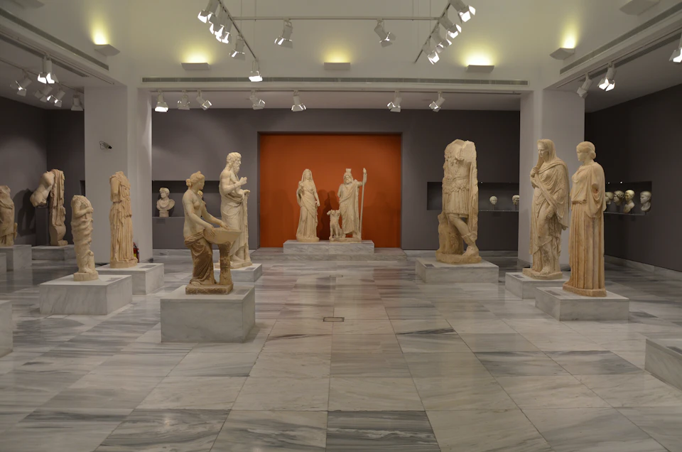 A museum gallery with marble floors displaying various ancient statues, including figures in togas and one of Athena holding a spear.