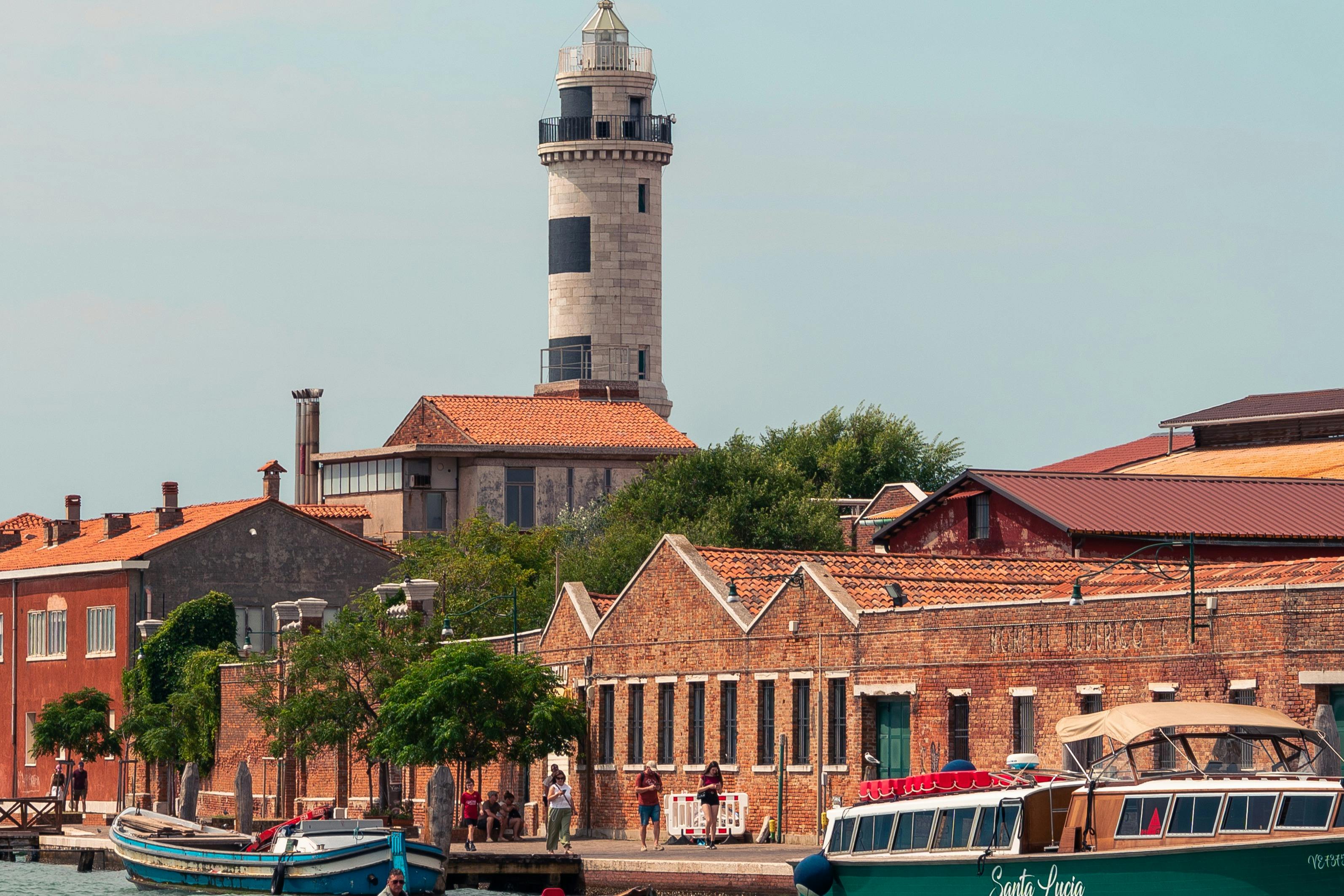 水辺のレンガ造りの建物の列の後ろに灯台があり、晴れた空の下で前景にボートが停泊しています。