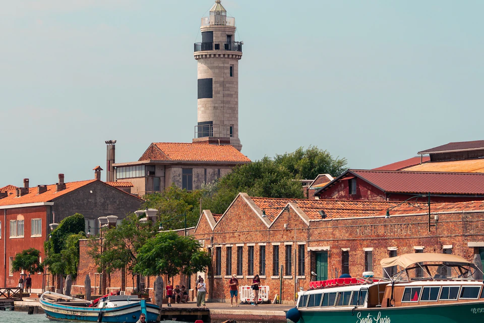 A lighthouse stands behind a row of brick buildings by the water, with boats docked in the foreground under a clear sky.