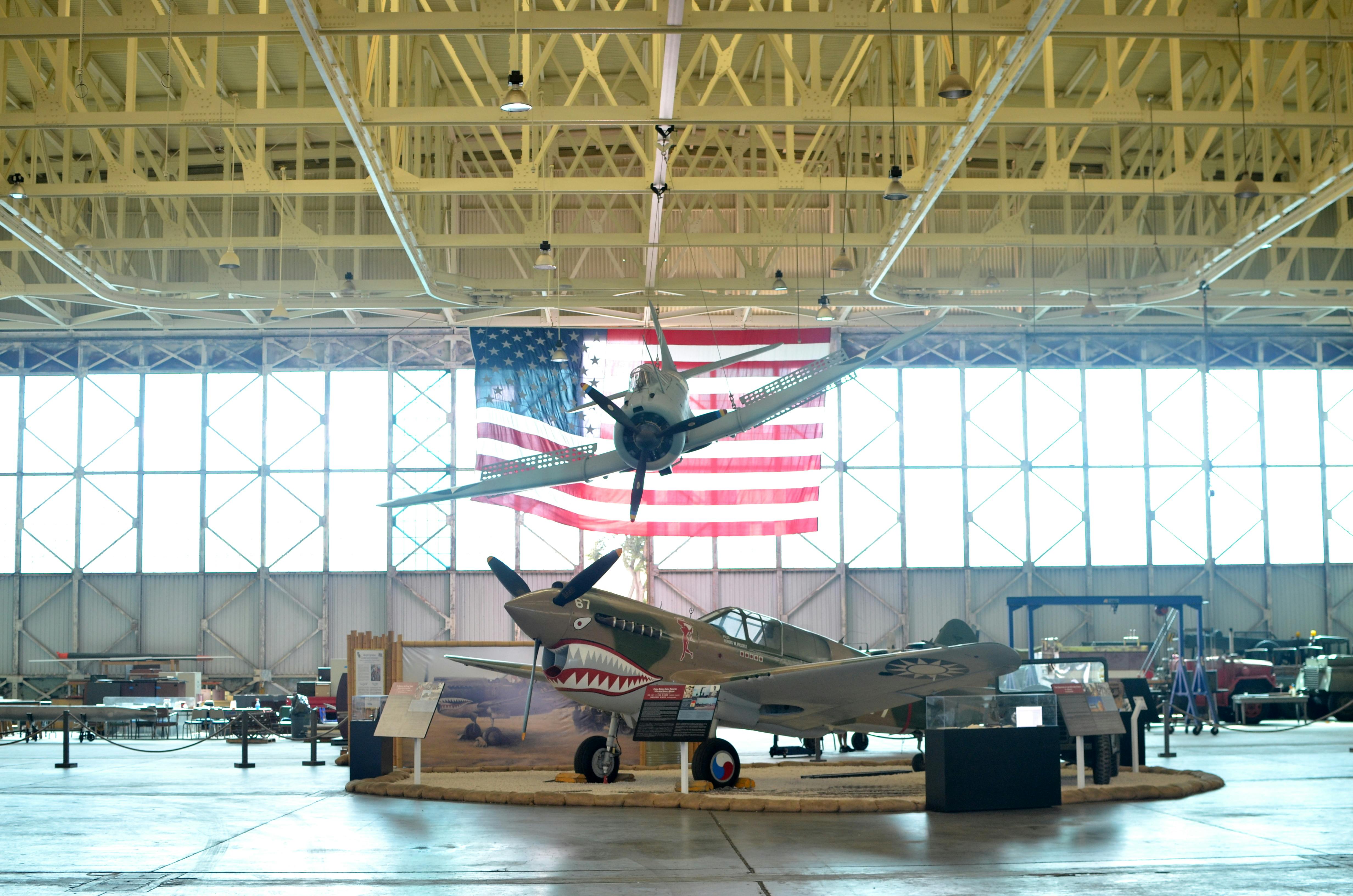 航空博物馆，陈列着一架二战战斗机，上面悬挂着另一架飞机；背景中悬挂着一面巨大的美国国旗。