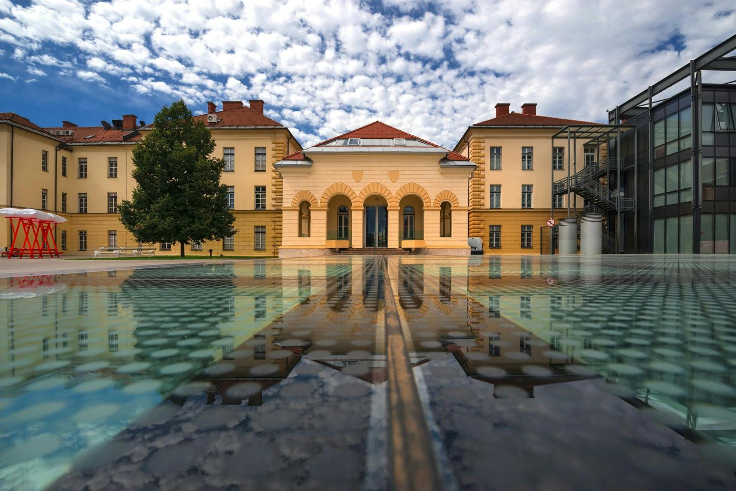 Symetrická historická budova s klenutými dveřmi se odráží v klidné vodní hladině pod částečně zataženou oblohou.