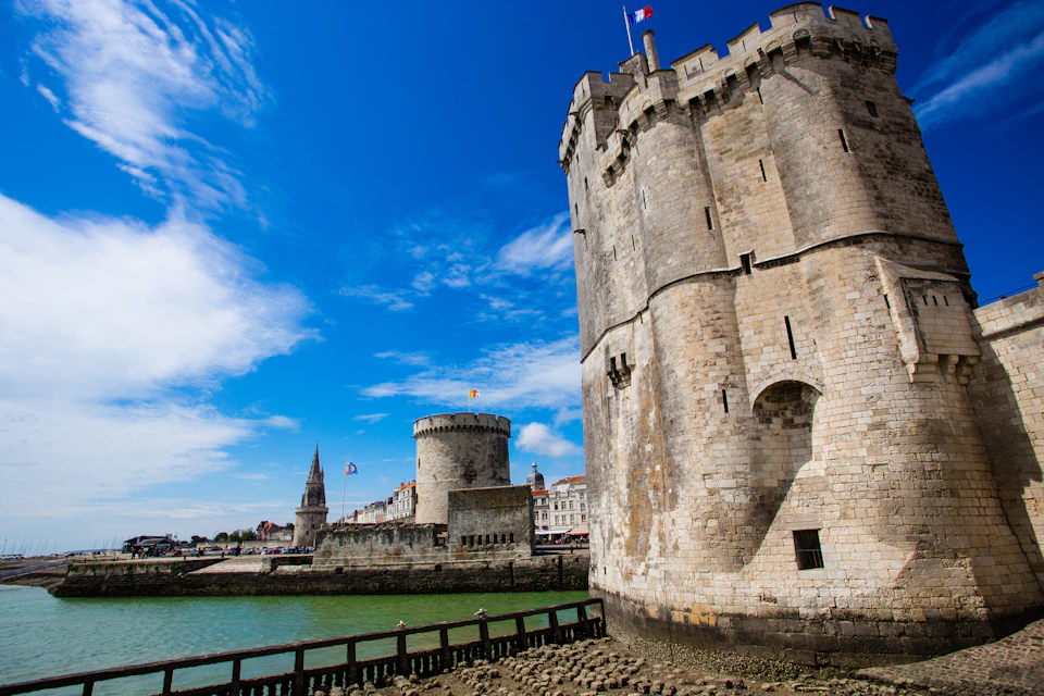 Two medieval towers by the water with a clear blue sky. Buildings and a church spire are visible in the background.