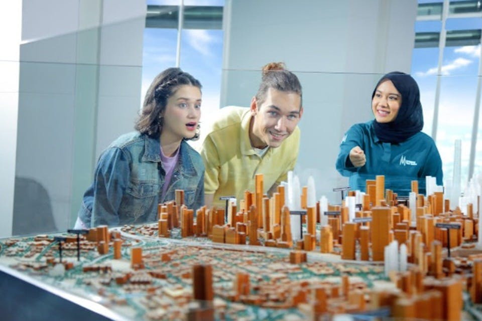 Dos personas examinan una maqueta detallada de una ciudad con edificios y calles, expuesta en una exposición cubierta de cristal.