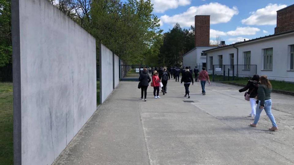People walking along a paved pathway lined with trees and a concrete wall under a partly cloudy sky.