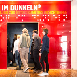 Människor går in i en mörk dörröppning under en röd skylt med texten "DIALOG IMDUNKELN ®" med ytterligare text på tyska.