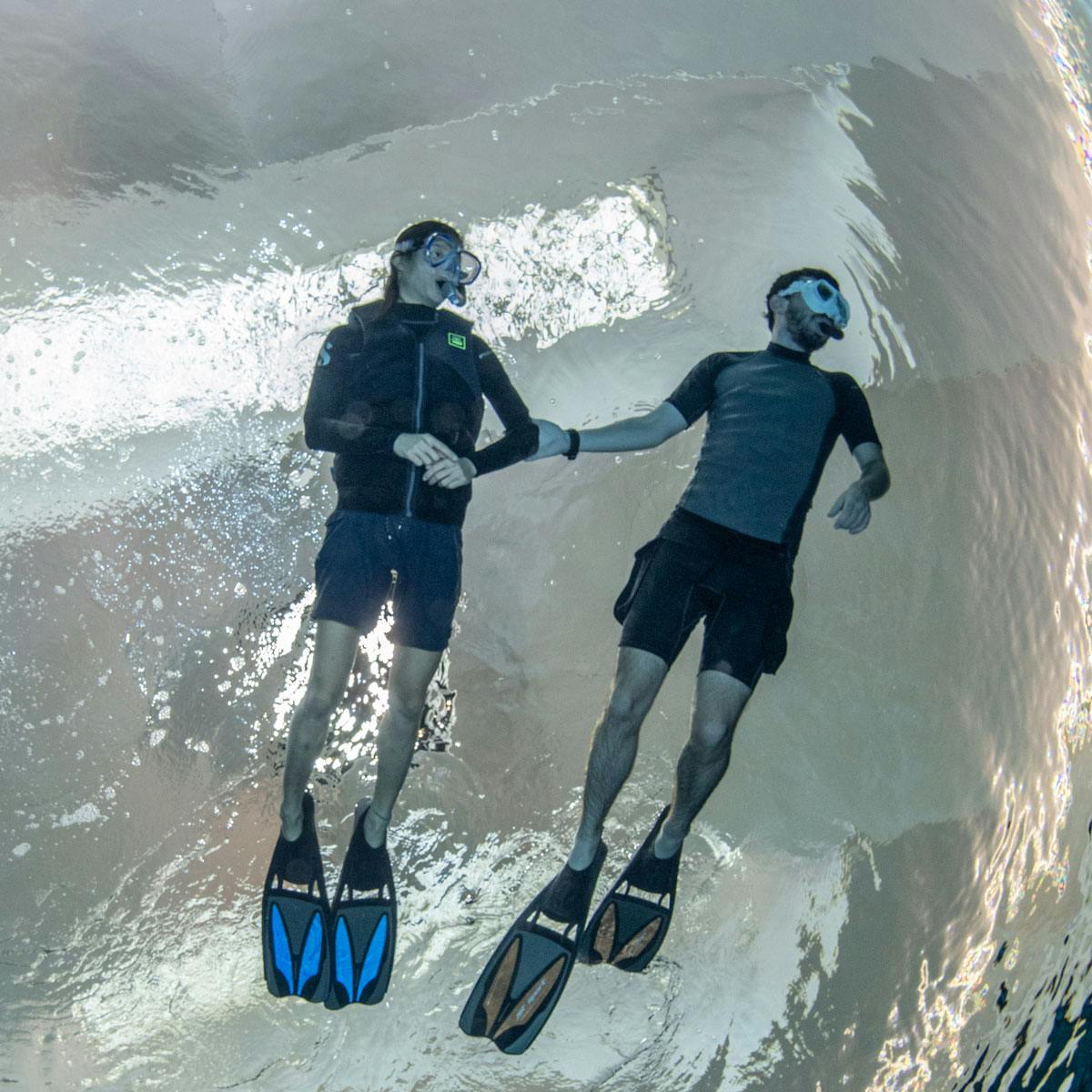 Δύο αναπνευστήρες κρατούν τα χέρια τους κάτω από το νερό, φορώντας αναπνευστήρες, μάσκες, βατραχοπέδιλα και στολές, που επιπλέουν κοντά στην επιφάνεια του νερού.