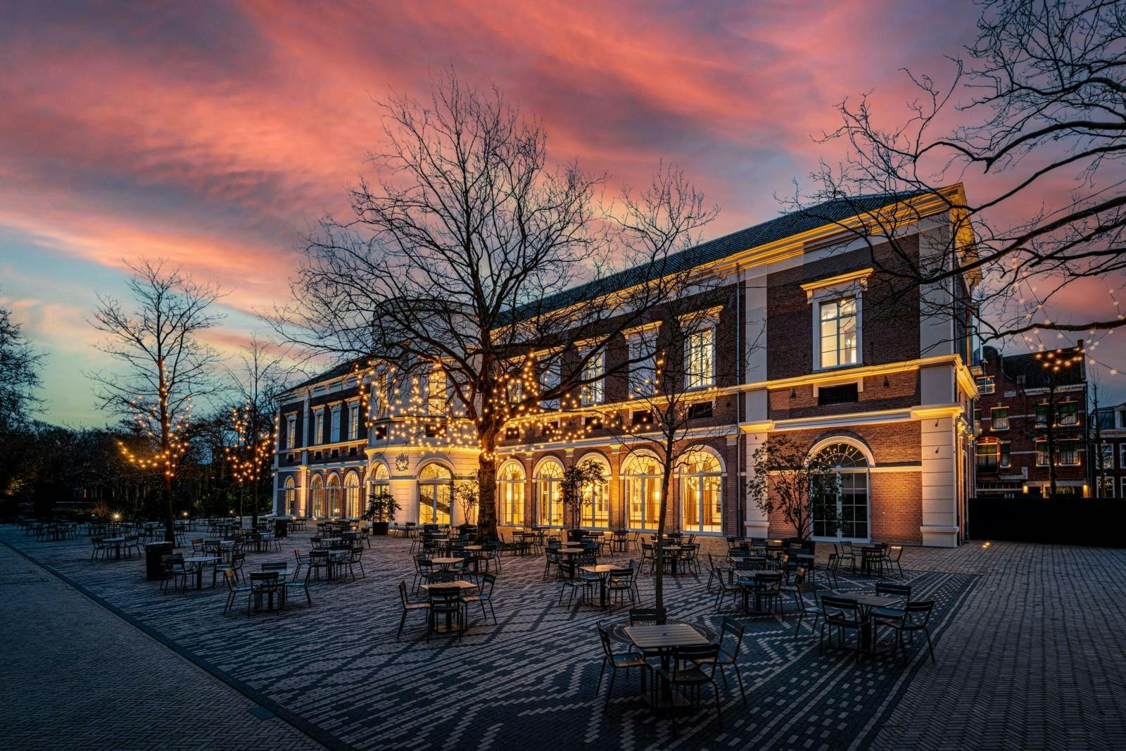 Een gebouw met verlichte ramen en een terras onder een boom met lampjes bij zonsondergang.