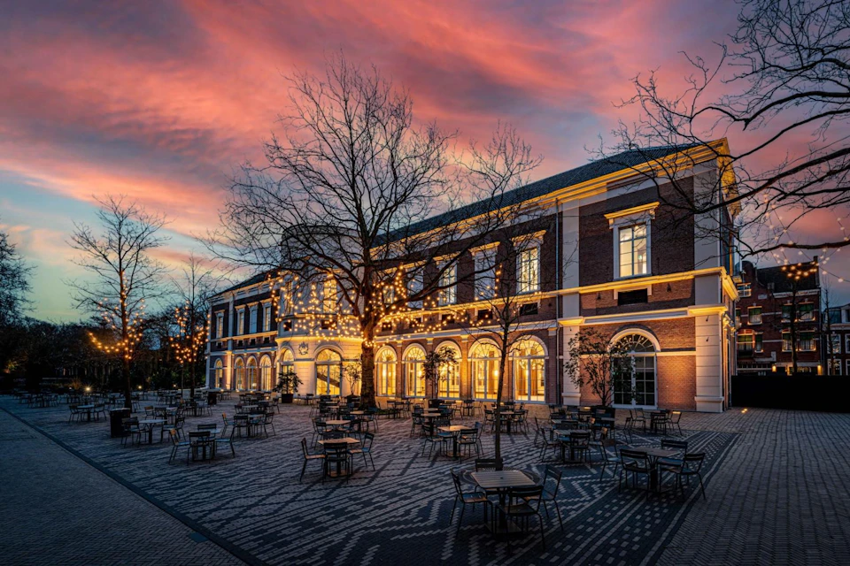 A building with illuminated windows and outdoor seating under a tree with lights at sunset.