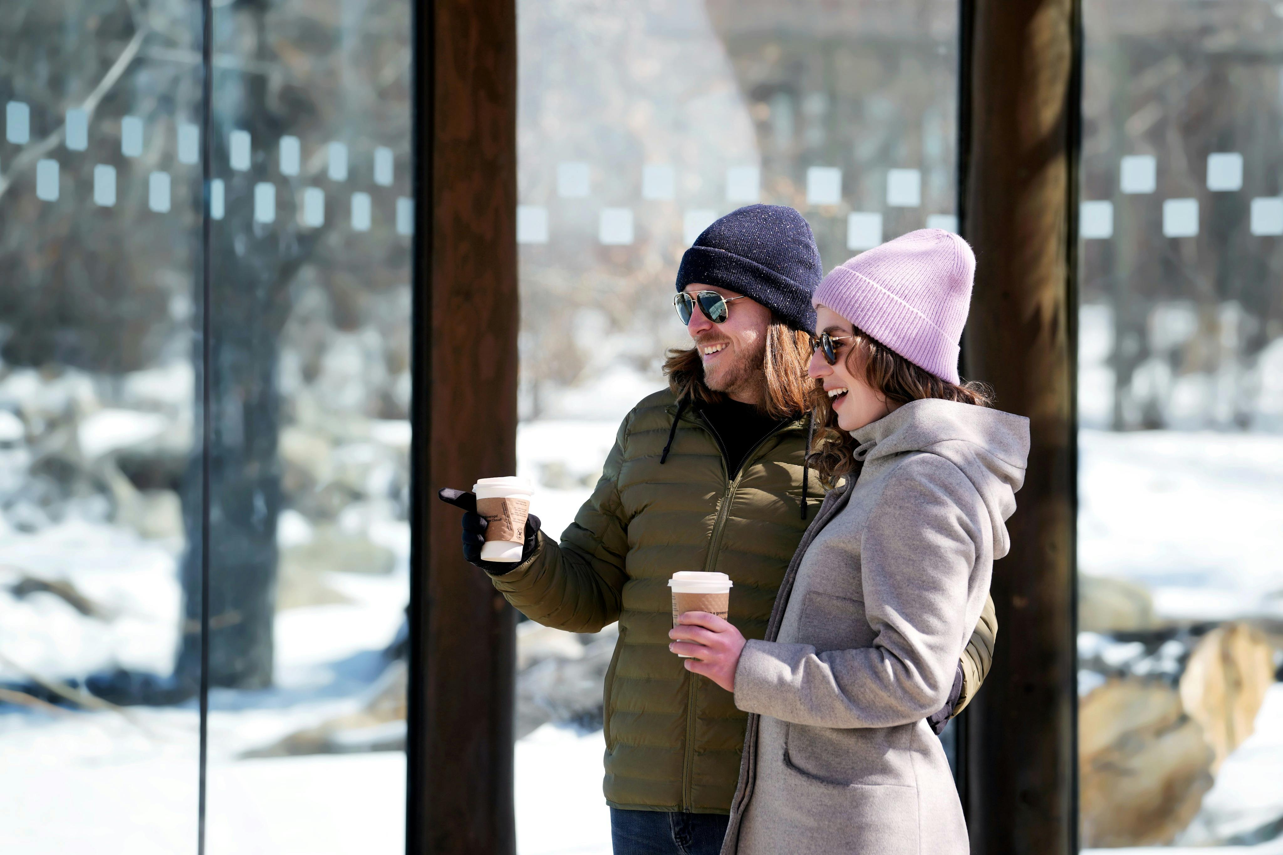 겨울옷을 입은 두 사람이 각각 커피잔을 들고 미소를 짓고 눈과 나무를 배경으로 야외에 서 있습니다.
