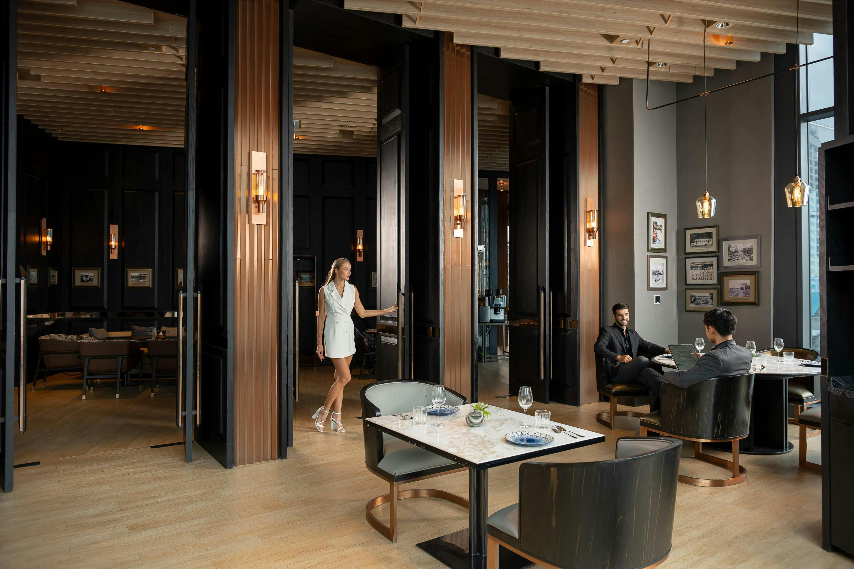 A modern dining area with people seated at tables and a woman walking, featuring stylish decor and pendant lighting.