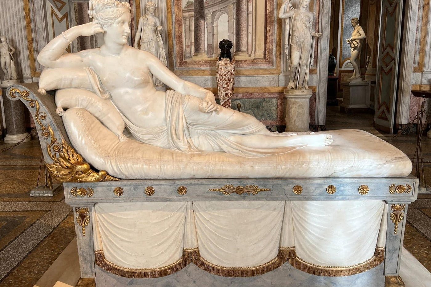 Una statua in marmo di una figura sdraiata in un ambiente classico, circondata da decorazioni e elementi architettonici decorati.