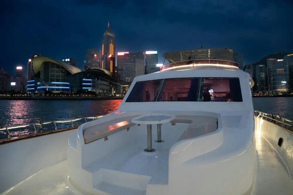 A yacht deck with seating and a table, with a city skyline illuminated against a night sky in the background.