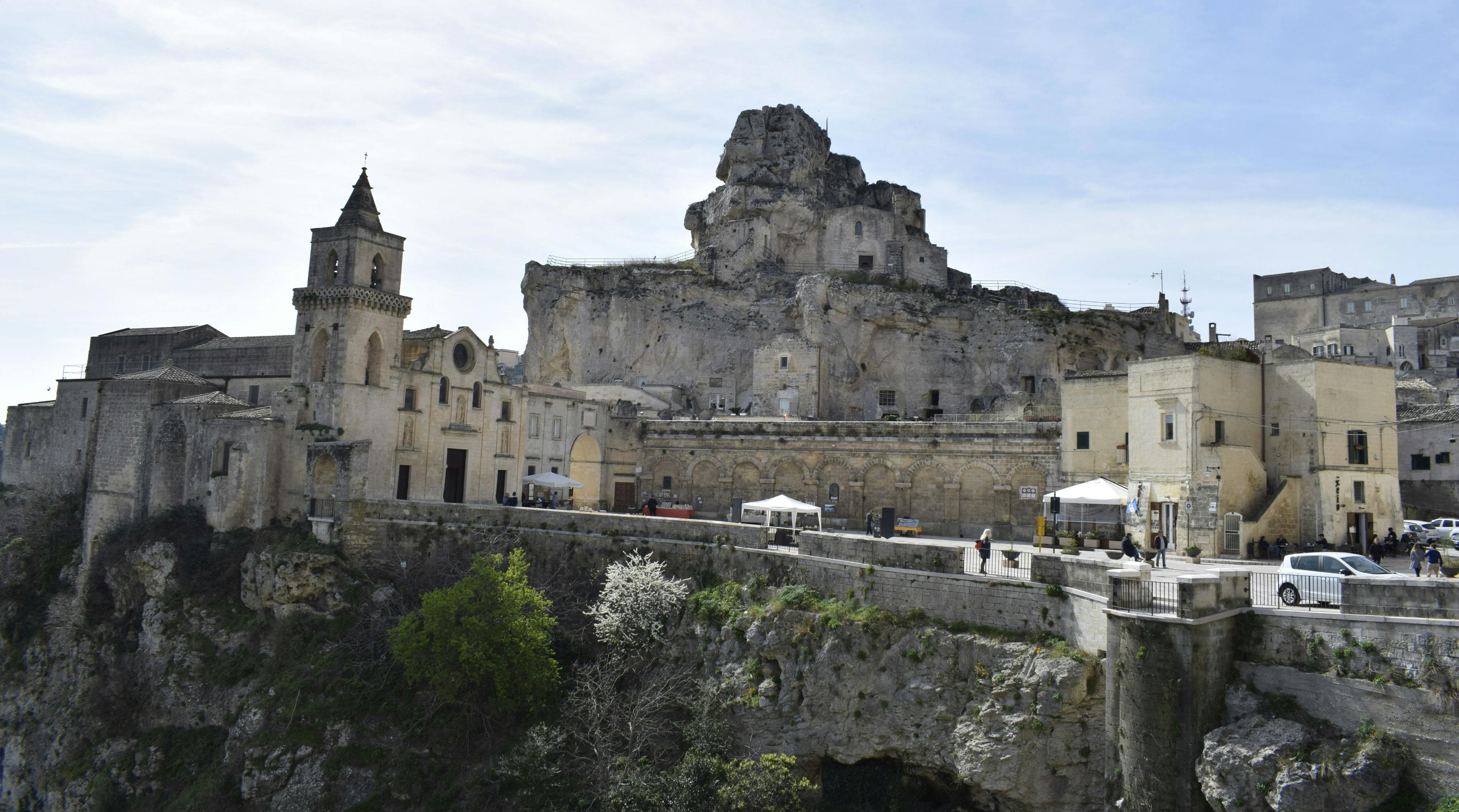 Ένας ιστορικός βραχώδης σχηματισμός με αρχαία κτίρια και ένα ψηλό καμπαναριό, με θέα σε μια βαθιά, δεντρόφυτη χαράδρα κάτω από έναν συννεφιασμένο ουρανό.
